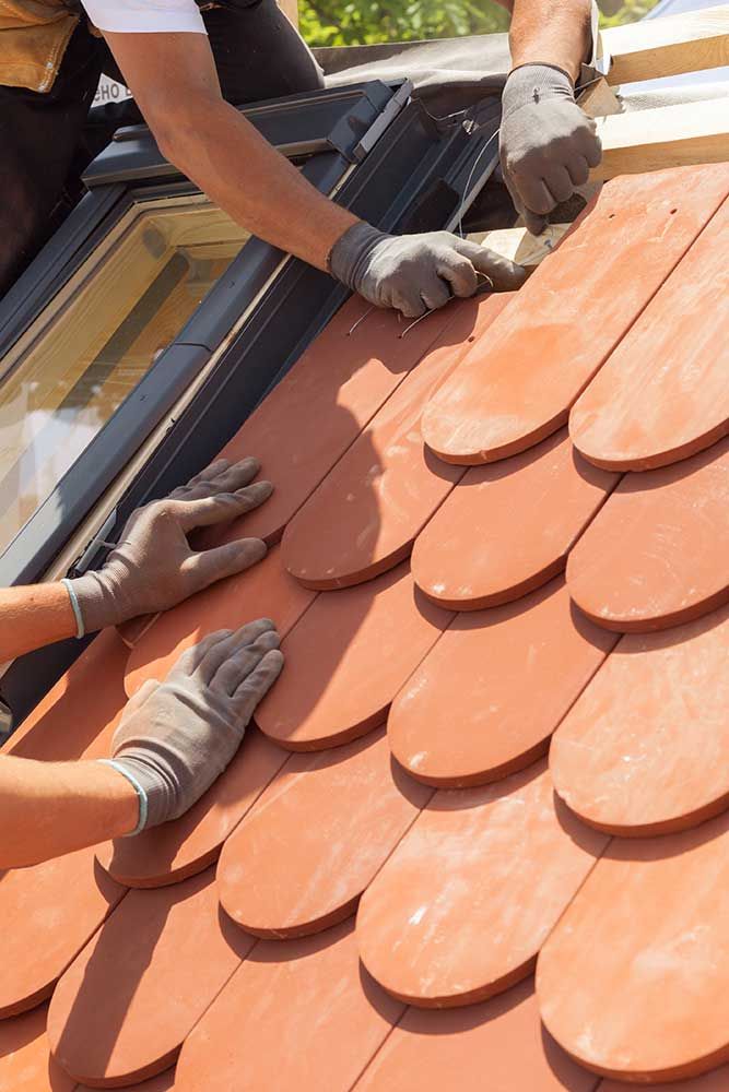 Hände des Dachdeckers, der Fliesen auf das Dach legt. Installieren von natürlichen roten Fliesen. Dach mit Mansardfenstern.